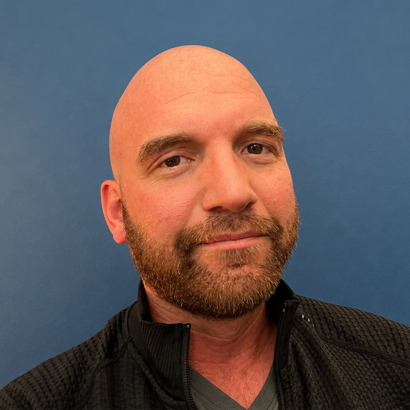 Close-up portrait of Caleb Zahnd, bald with a full beard, wearing a dark jacket against a plain blue background.